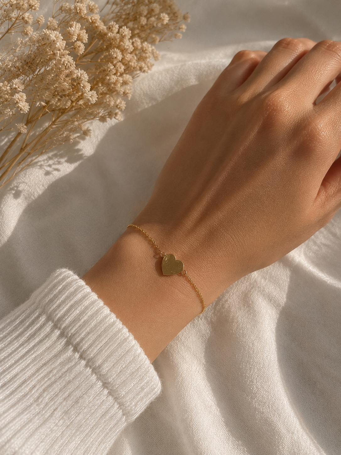 Gold-toned heart charm chain bracelet worn on a wrist beside dried flowers