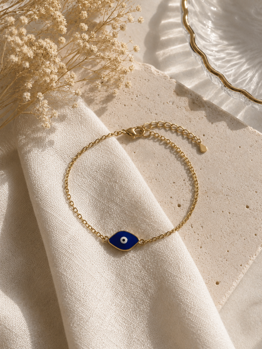 Gold-toned chain bracelet with a blue enamel marquise evil eye charm on cream fabric