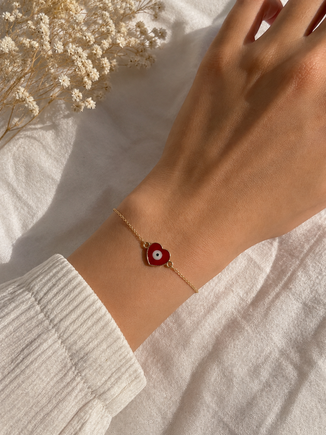 Red enamel heart evil eye bracelet worn on a wrist beside dried flowers