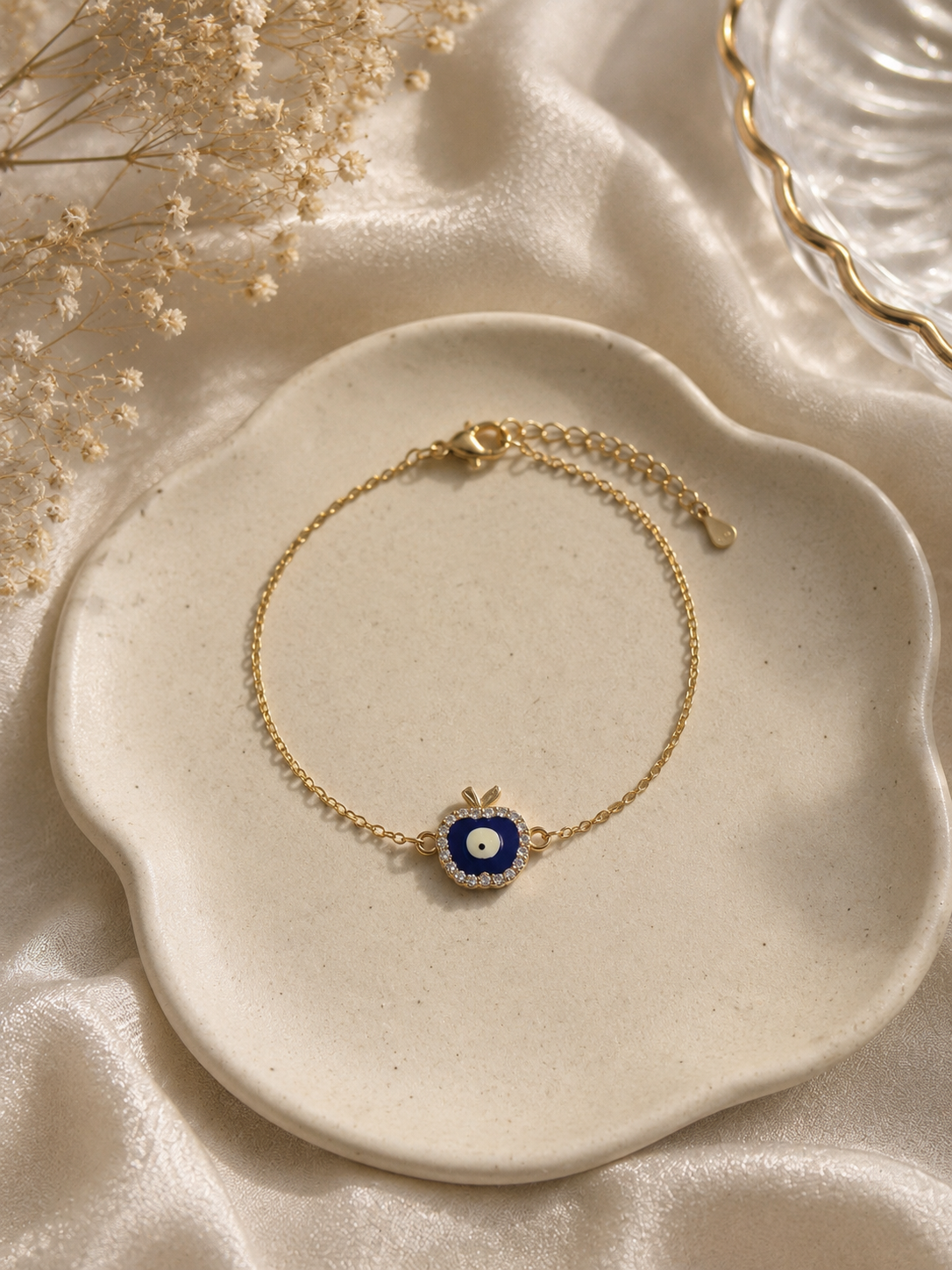 Gold-toned bracelet with a blue enamel apple evil eye charm on a scalloped cream dish