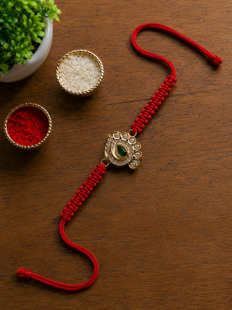 Gold paisley kundan rakhi with a green centre stone on a red thread, on wood with puja bowls