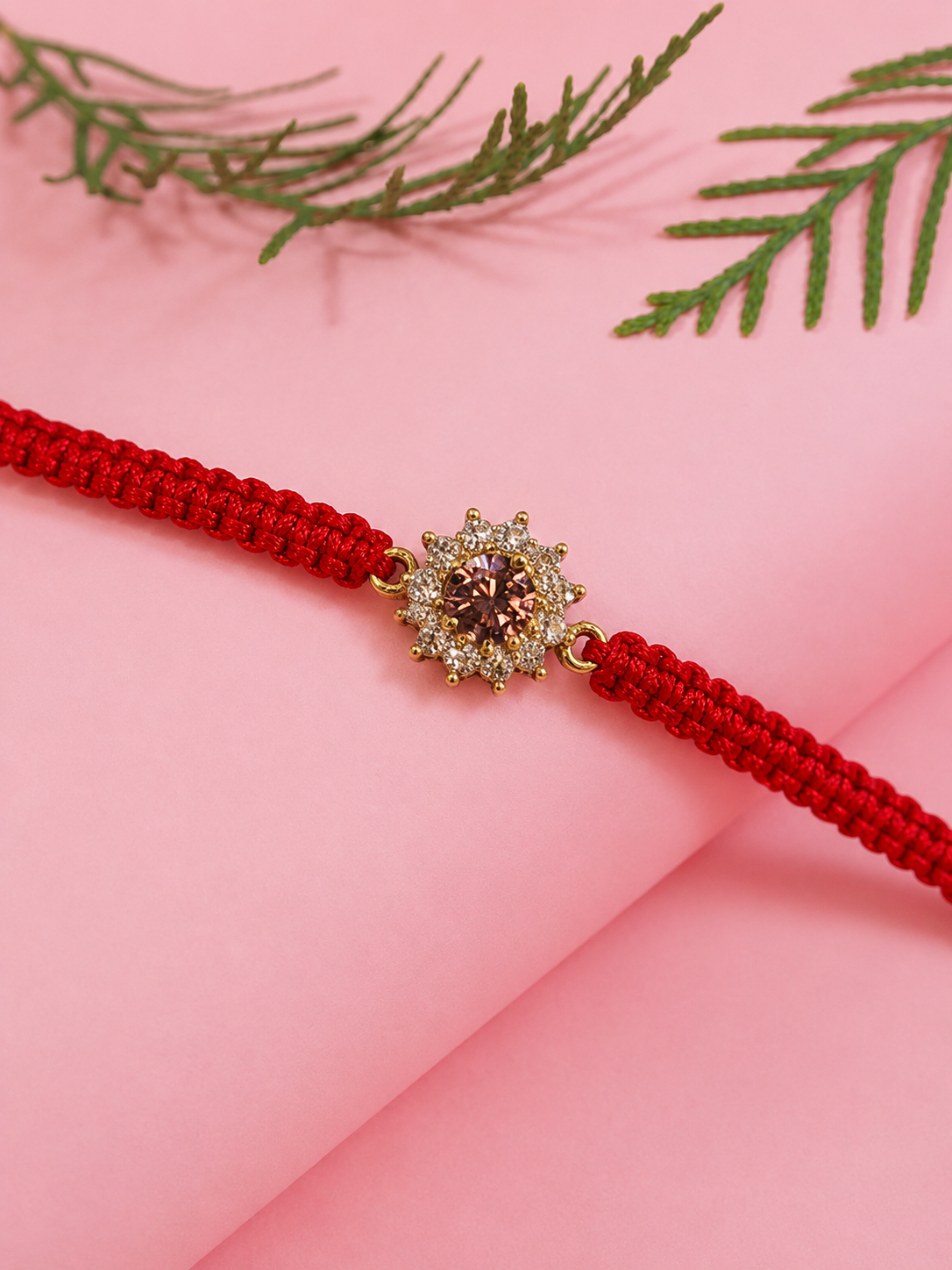 Gold floral rakhi with pink stones on a red thread on a pink backdrop