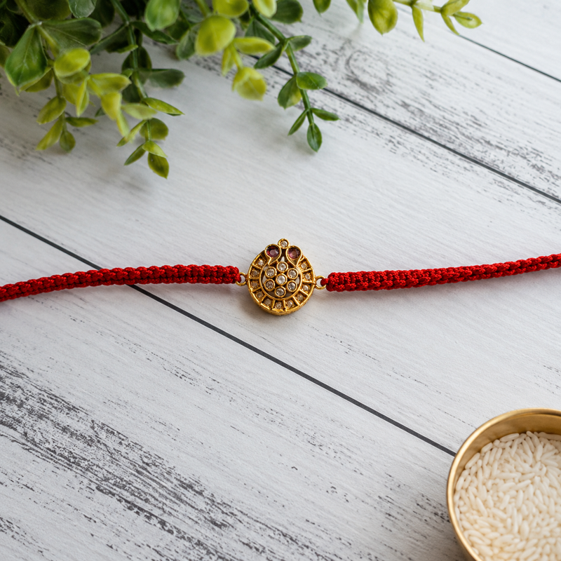 Gold antique round disc rakhi on a red thread on a white wooden surface with greenery