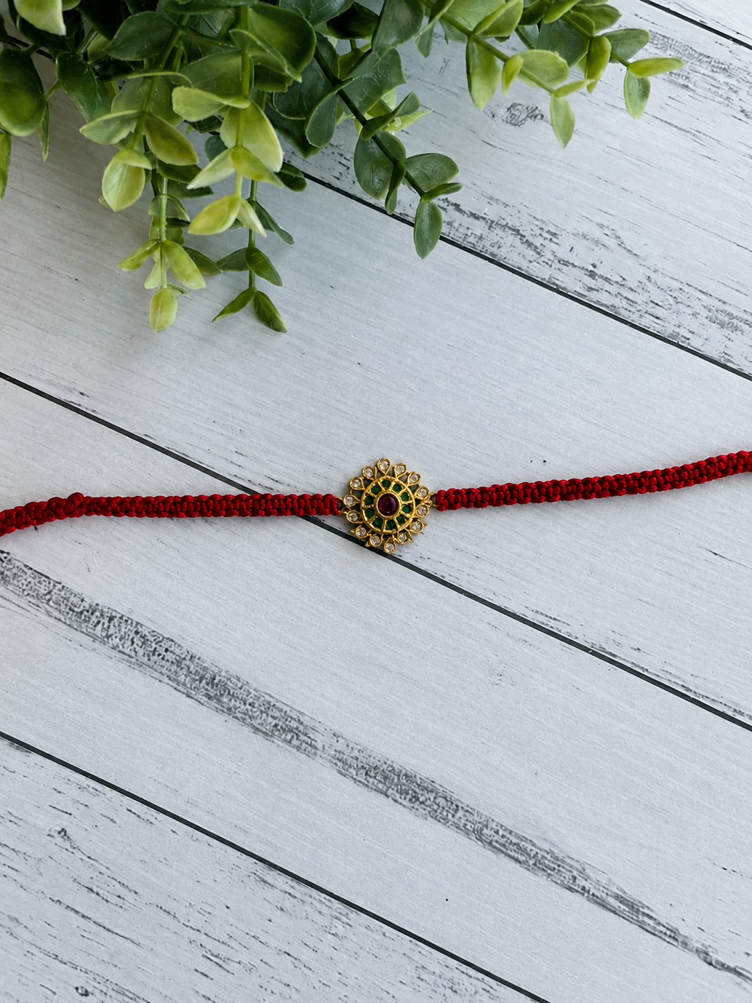 Gold kemp blossom rakhi with a green centre and red petals on a red thread on white wood