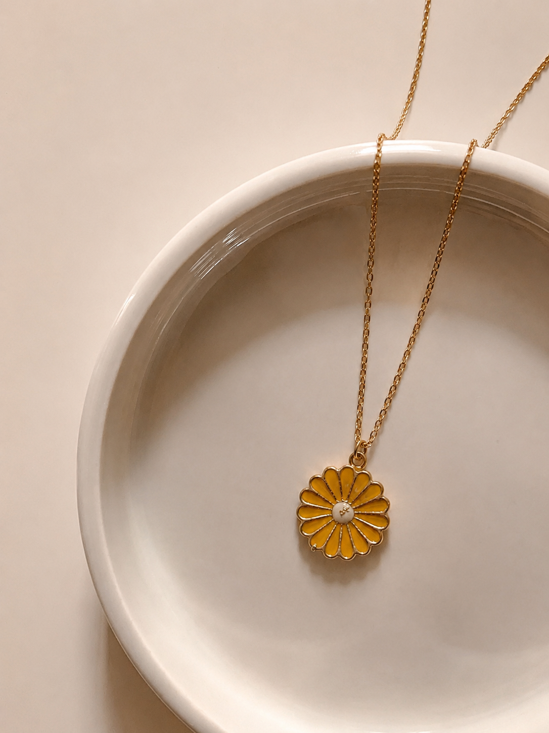 Gold-toned rope chain with a mustard-yellow enamel daisy pendant on a white plate