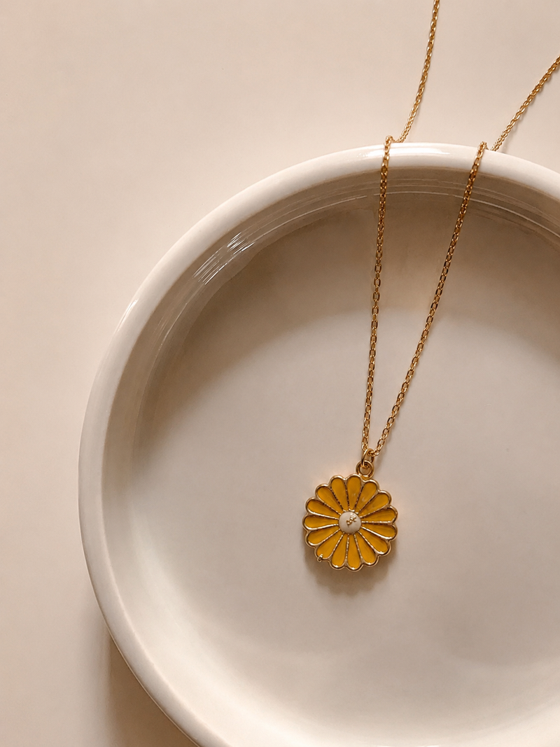 Gold-toned rope chain with a mustard-yellow enamel daisy pendant on a white plate