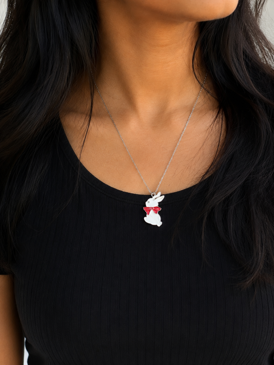 White enamel bunny pendant with red bow worn on a chain over a black top