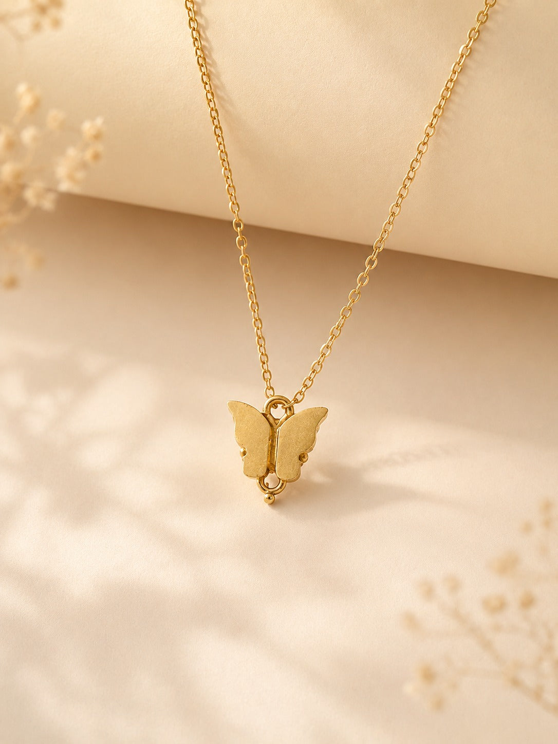 Gold-toned butterfly pendant on a fine chain resting on a cream surface with dried flowers