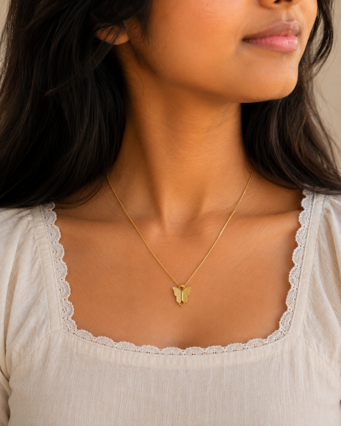Close view of the gold-toned butterfly pendant worn on a dainty chain over a cream top