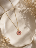 Pink enamel daisy pendant on a gold-toned chain styled on a cream dish with dried flowers