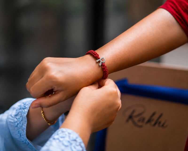 Silver initial R rakhi on a red thread being tied on a wrist