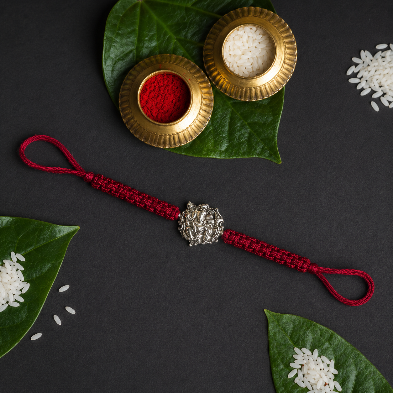 Silver Ganesha bead rakhi on a looped red thread on a dark backdrop with betel leaves